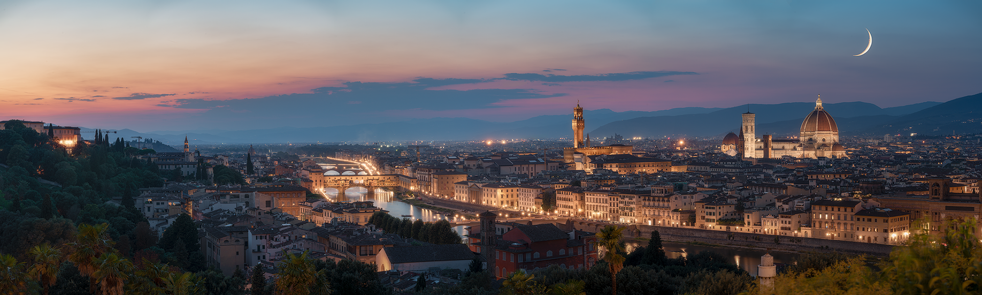 Firenze vista dall’alto sotto un cielo sereno, con la luna crescente che aggiunge un tocco magico alla scena. Il Duomo, Palazzo Vecchio e il Ponte Vecchio sono illuminati, risaltando il fascino senza tempo della città.