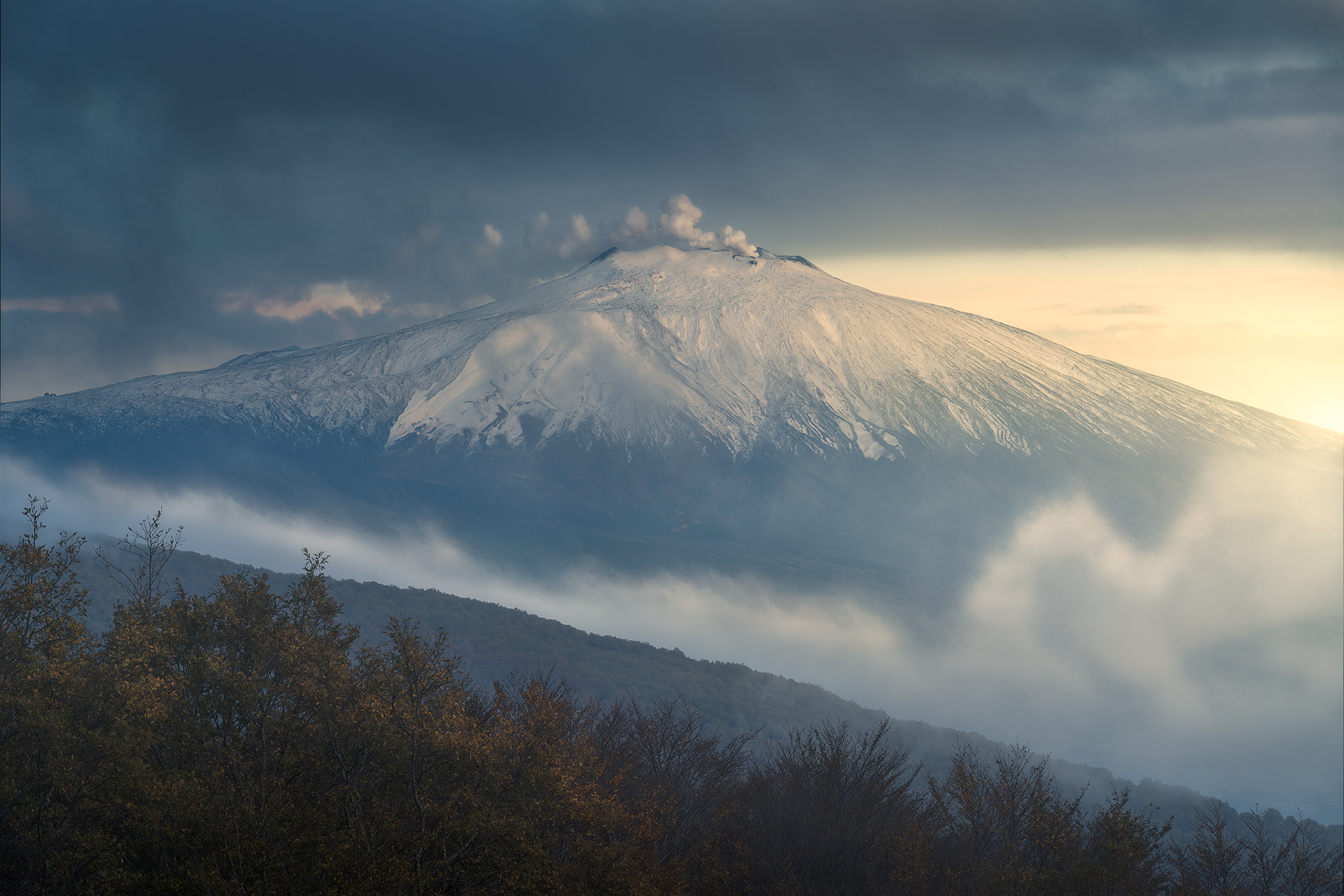 Vista dell’Etna avvolto dalle nuvole Il vulcano si staglia imponente con il suo profilo innevato, mentre nuvole minacciose si avvolgono attorno alle sue pendici. Un paesaggio drammatico che trasmette tutta la potenza e la maestosità dell’Etna.