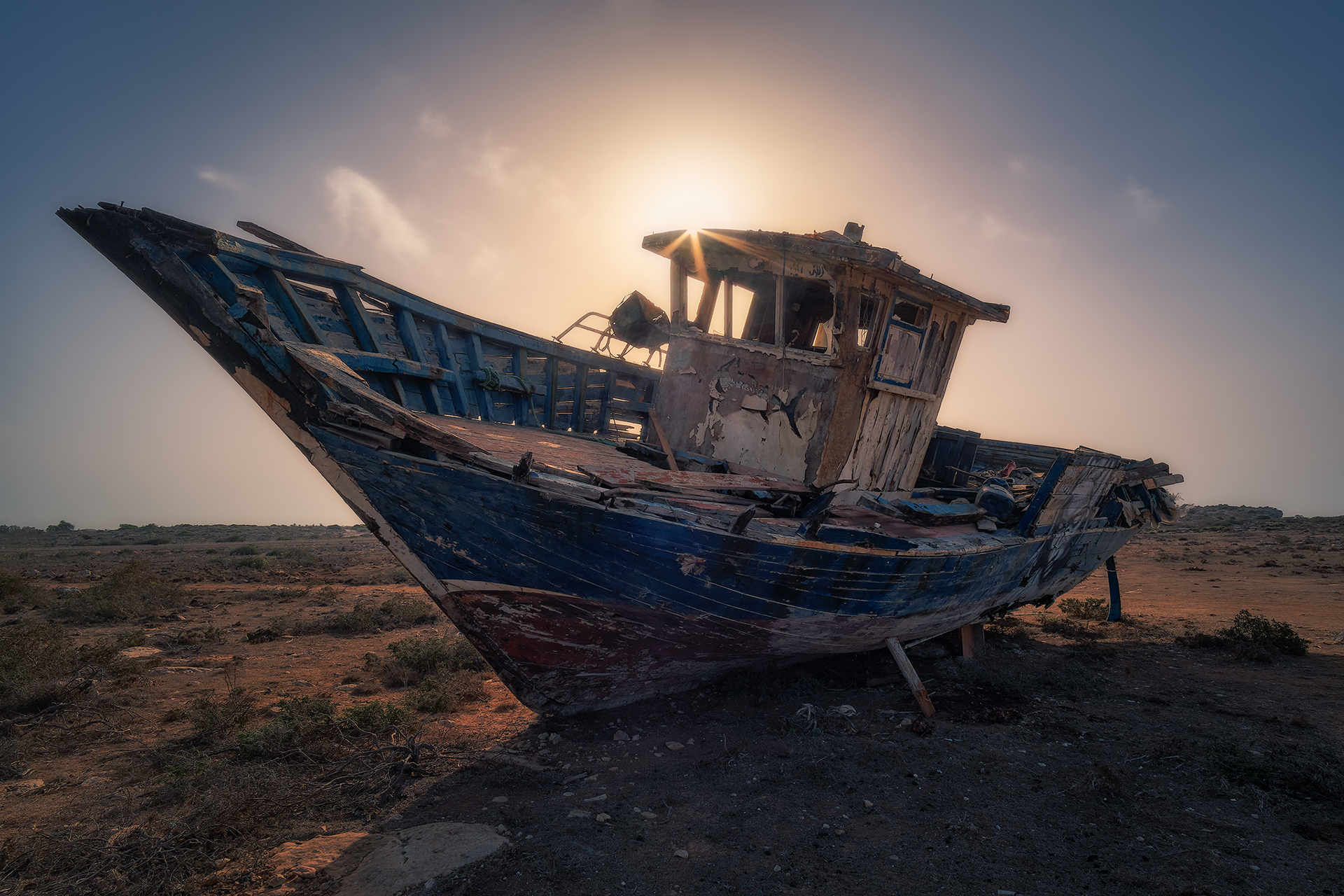 Un relitto di barca usata dai migranti giace abbandonato sulla costa di Lampedusa, testimone silenzioso di viaggi drammatici nel Mediterraneo. Il sole alle spalle crea un effetto drammatico che enfatizza il significato simbolico di questa immagine.