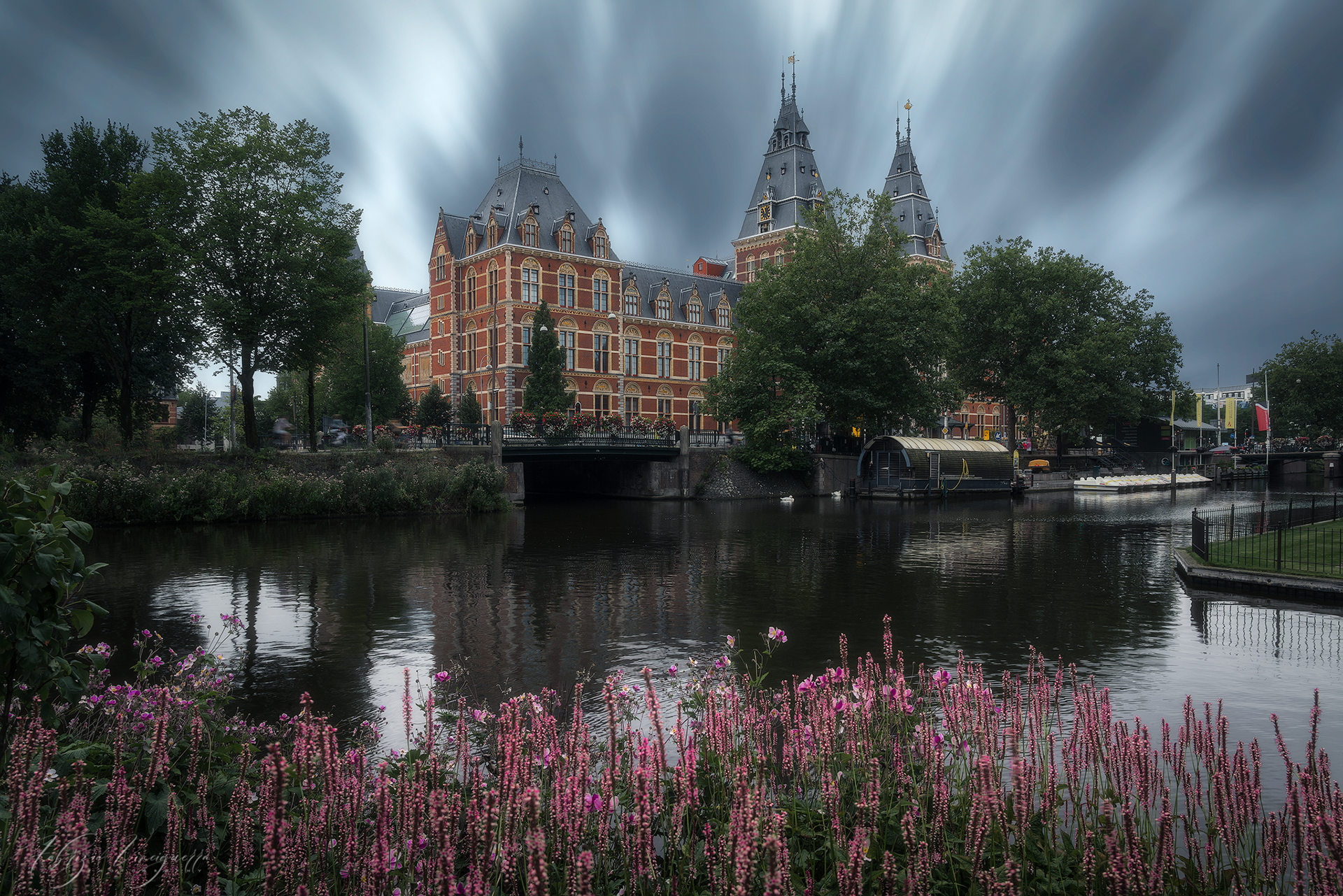 Edificio storico e canale fiorito – Un equilibrio perfetto tra architettura e natura, con il rosso acceso dei fiori che aggiunge un tocco di colore alla scena.