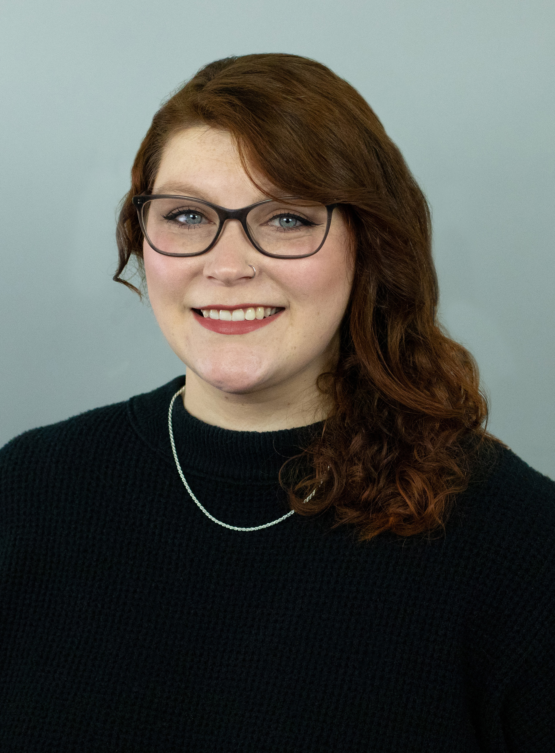 Professional headshot of Shannon Cowie, Senior Graphic Designer. Shannon is wearing a silver chain necklace, black-framed glasses and a black sweater.