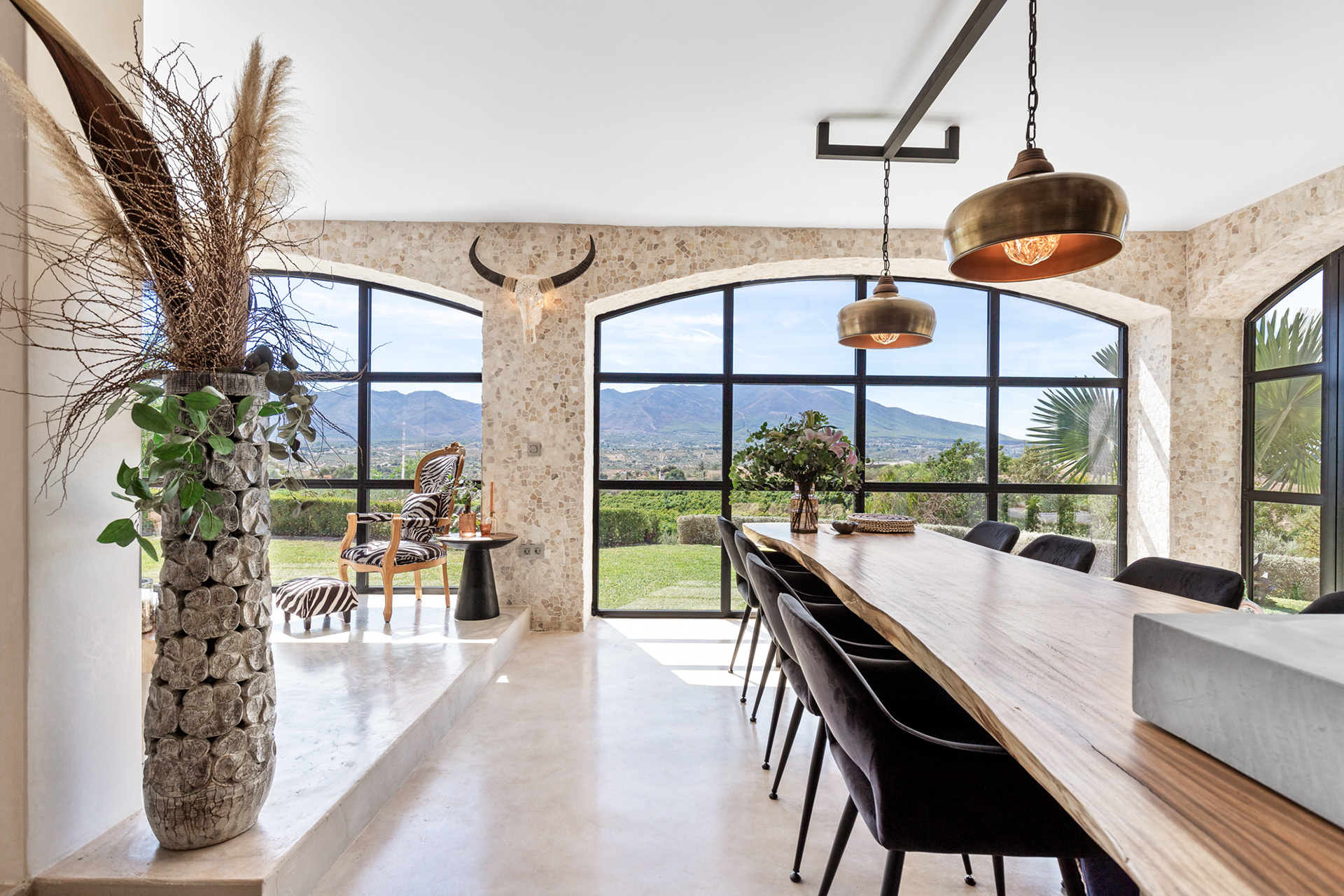 Luxury dining area photographed with professional real estate photography, featuring expansive arched windows, natural textures, designer lighting, and panoramic mountain views. Crafted to highlight premium interiors, architectural details, and the lifestyle appeal of high-end properties and villa's.