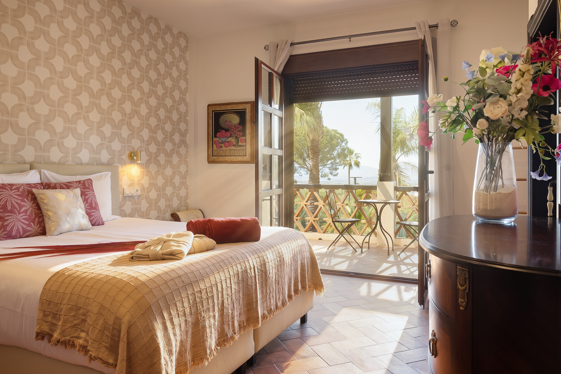 Bright and inviting bedroom captured with professional real estate photography, featuring warm natural light, elegant styling, retro-patterned wallpaper, and an open balcony with palm tree and landscape views. Ideal imagery to showcase hospitality spaces, vacation rentals, and property interiors with a welcoming atmosphere.