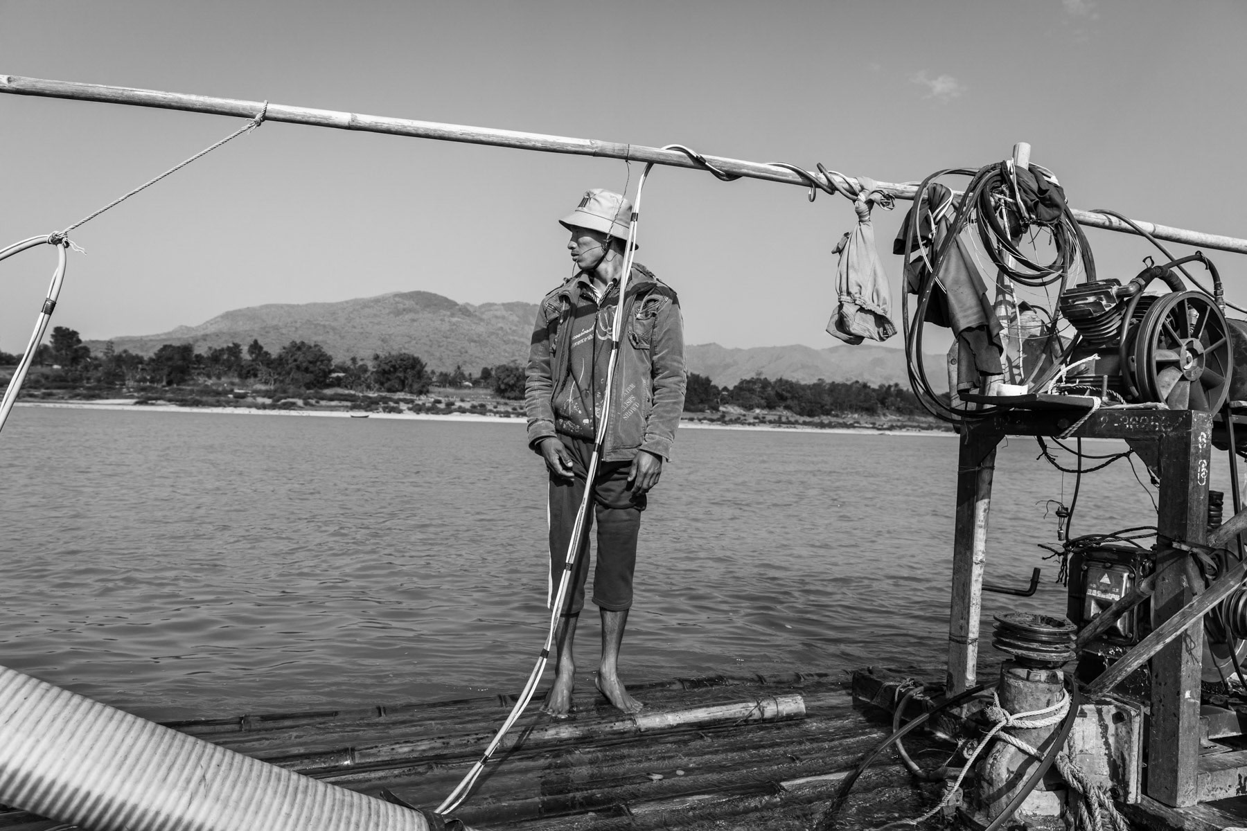 The helper and second diver waiting and controlling the compressor on the raft while his colleague is under water collecting soil and sediment that will be separated on the raft in order to recover gold residue. Irrawaddy River north of Myitkyina, Kachin State, Myanmar, February 2020