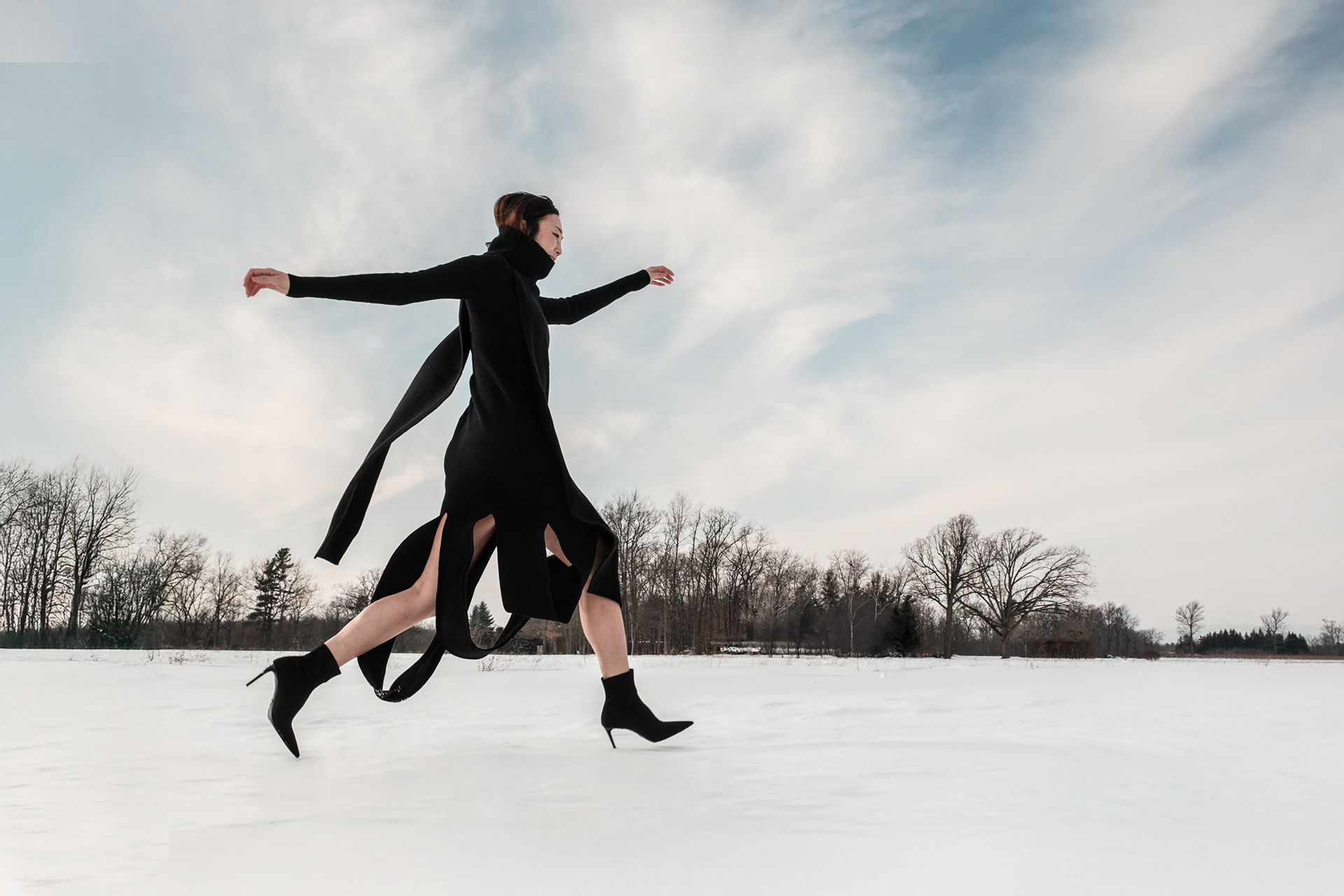 Person in a long black coat and high-heeled boots running across a snowy field with arms outstretched under a cloudy sky.