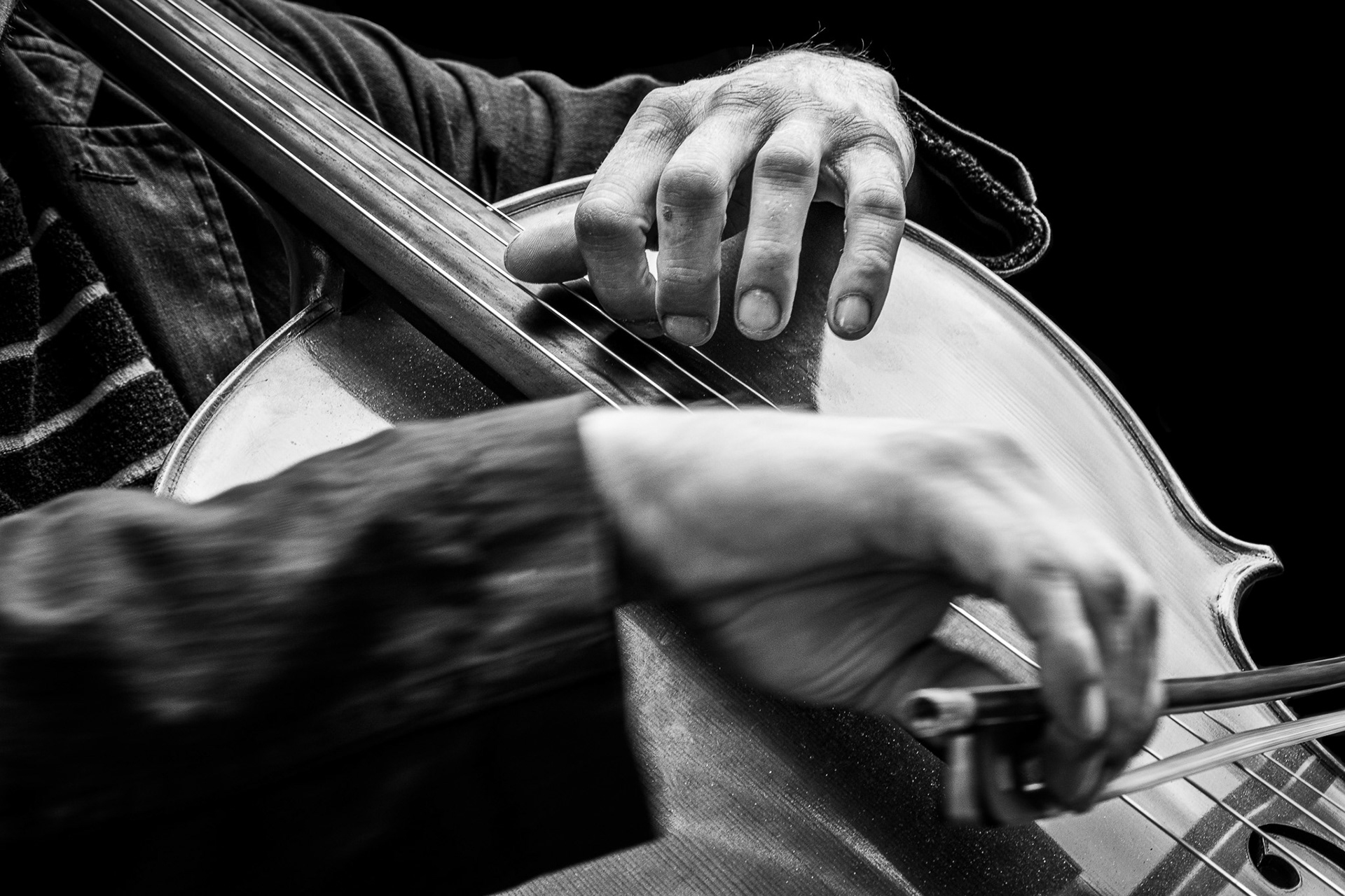 Een zwart‑wit close‑up van een cellospeler. De handen van de muzikant zijn zichtbaar: één hand drukt de snaren in op de hals van de cello, de andere beweegt de strijkstok over de snaren.