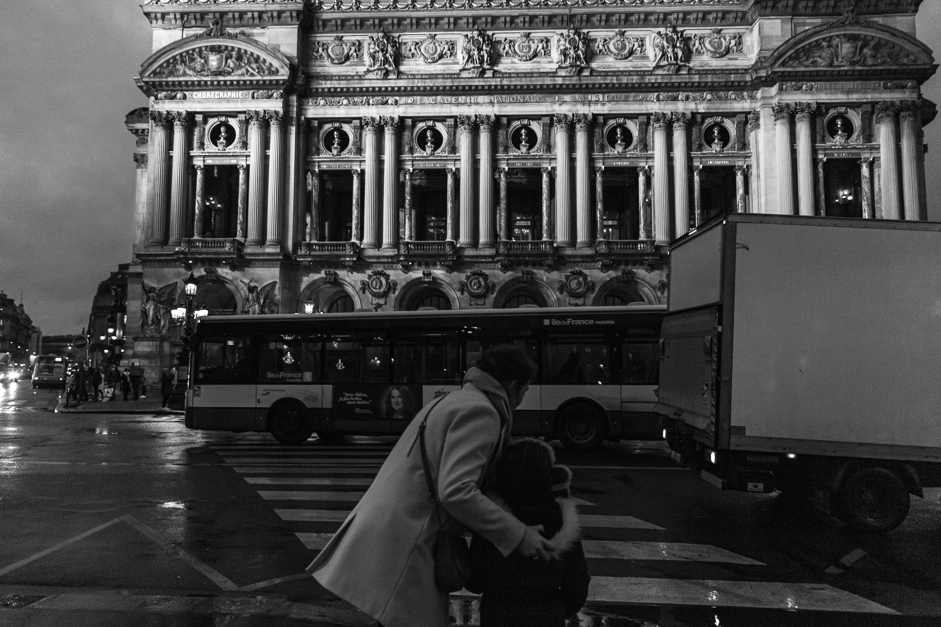 A mother holding her son gently by the hood as they wait to cross a street