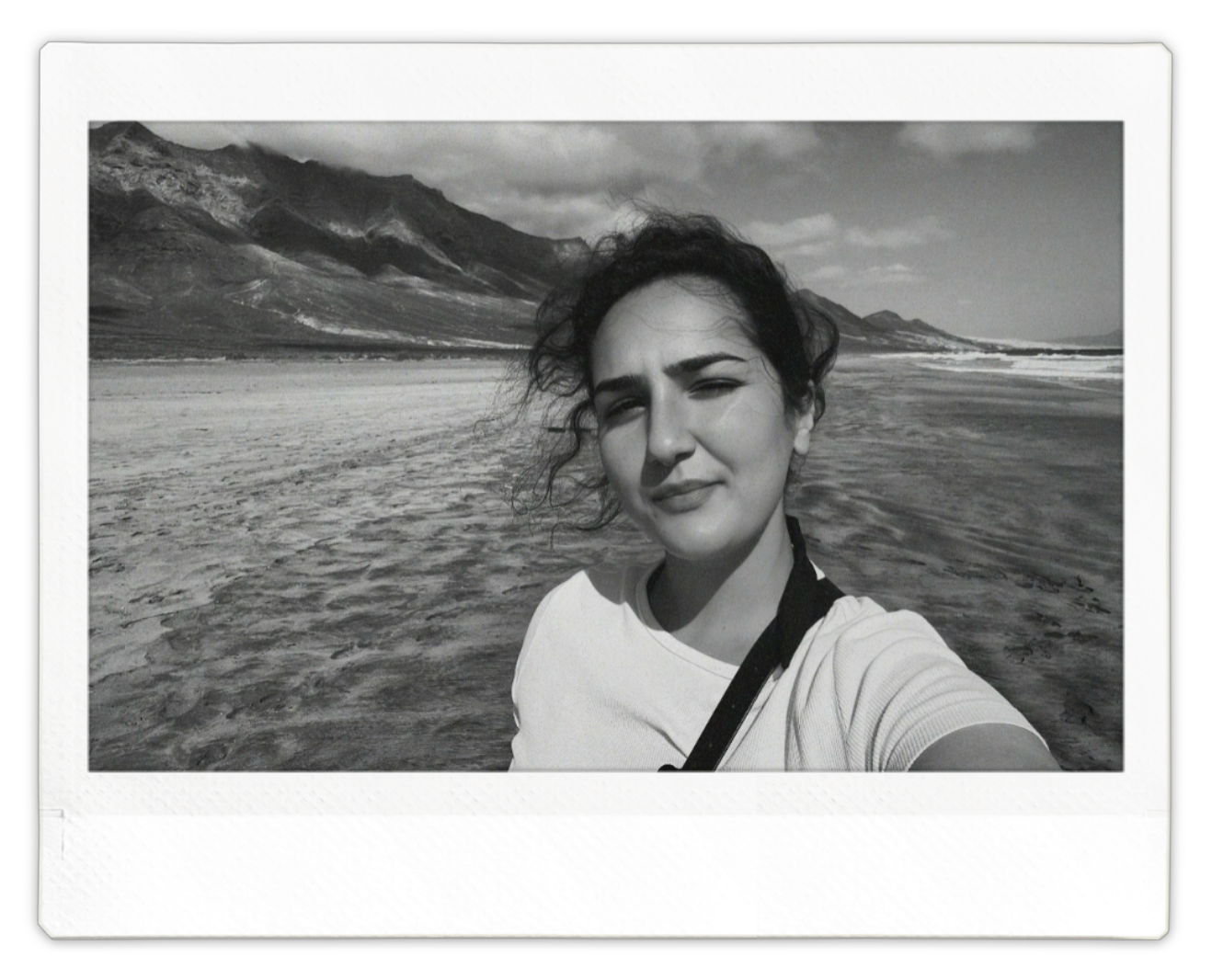 Black-and-white self-portrait of a woman standing in shallow ocean water, mountains in the background—analogue-style photography evoking solitude and awe.
