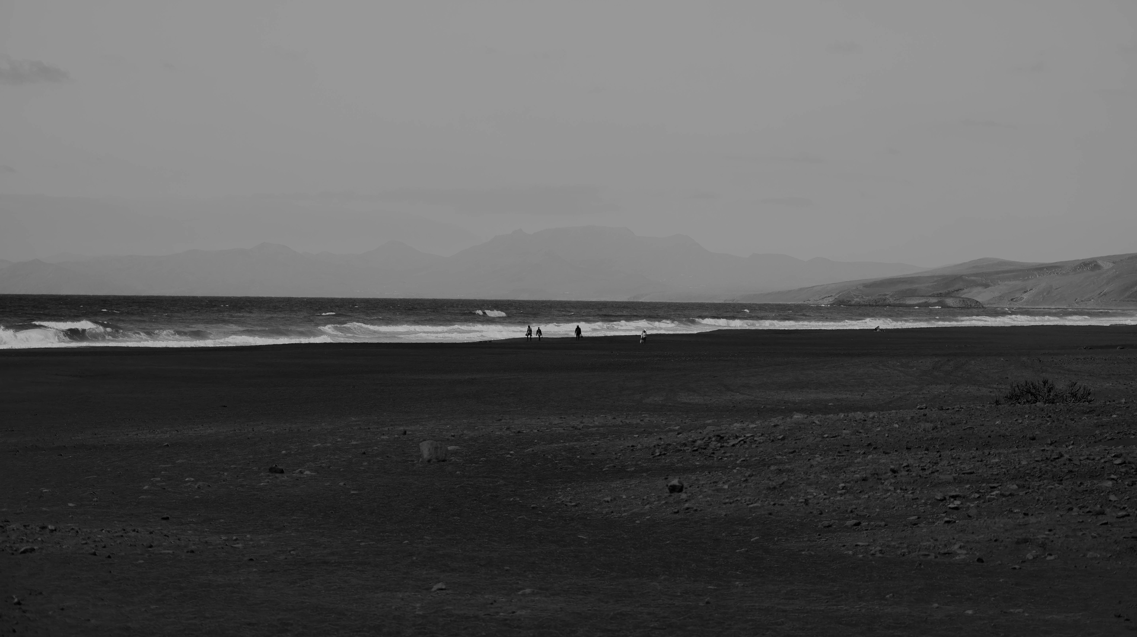 Black-and-white coastal landscape with mountains in the background and waves brushing a dark shoreline—minimalist and cinematic.