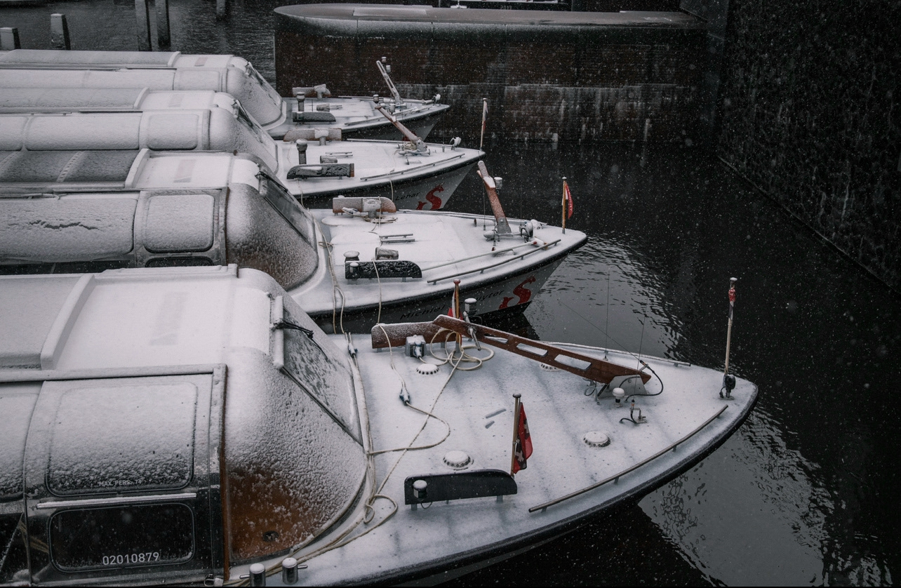 Angled view of multiple canal boats dusted in snow, reflected in dark winter waters—quiet moment captured during Amsterdam snowfall.