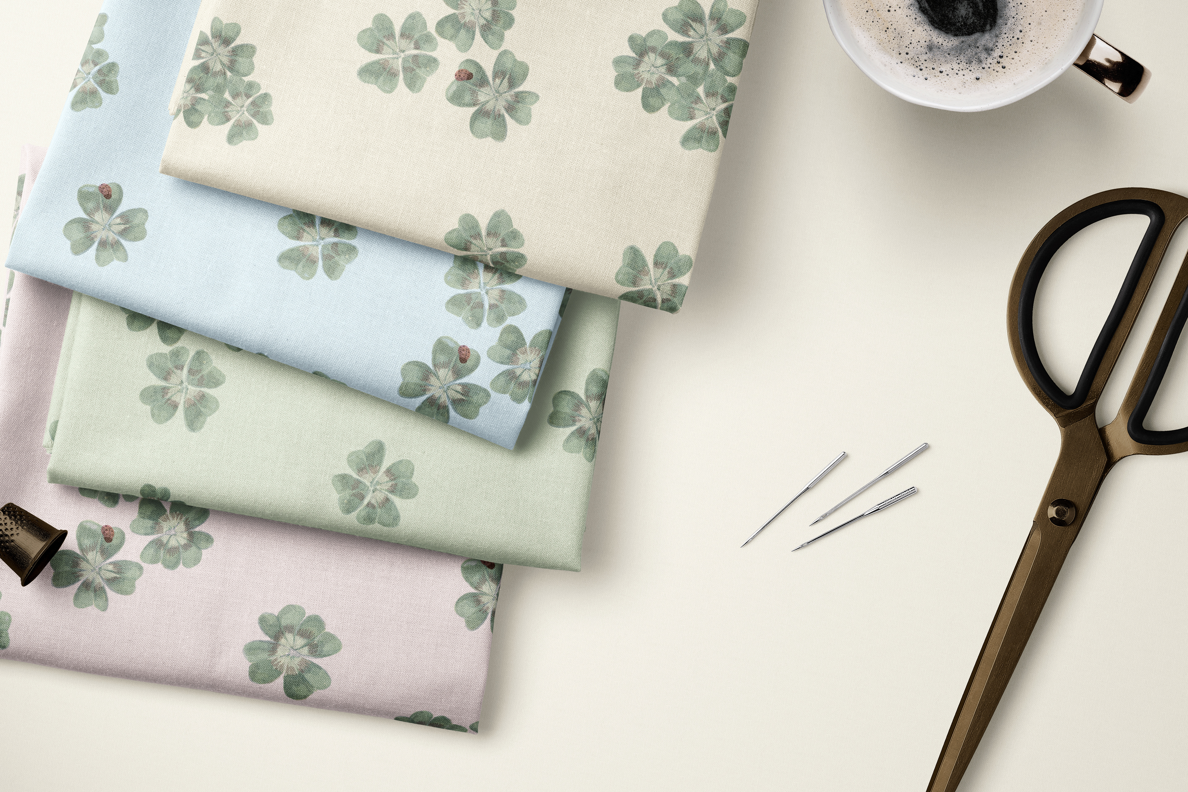 Flat lay of folded fabric swatches in Clover Scatter botanical print, shown in cream, sage green and blush pink colourways, with vintage scissors and sewing pins on a cream background