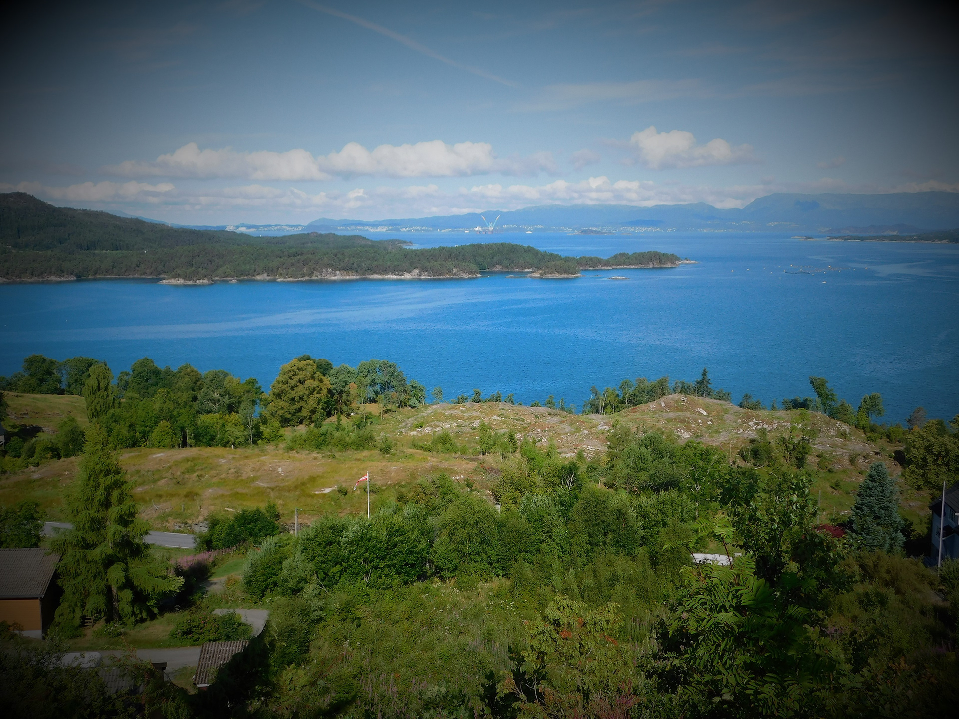 Mot vest ser du Sydnessundet (nærast), Klosterfjorden (til høgre), og Stordafjorden (bak). Legg merke til oljeplattforma i bakgrunnen. Juli 2018.