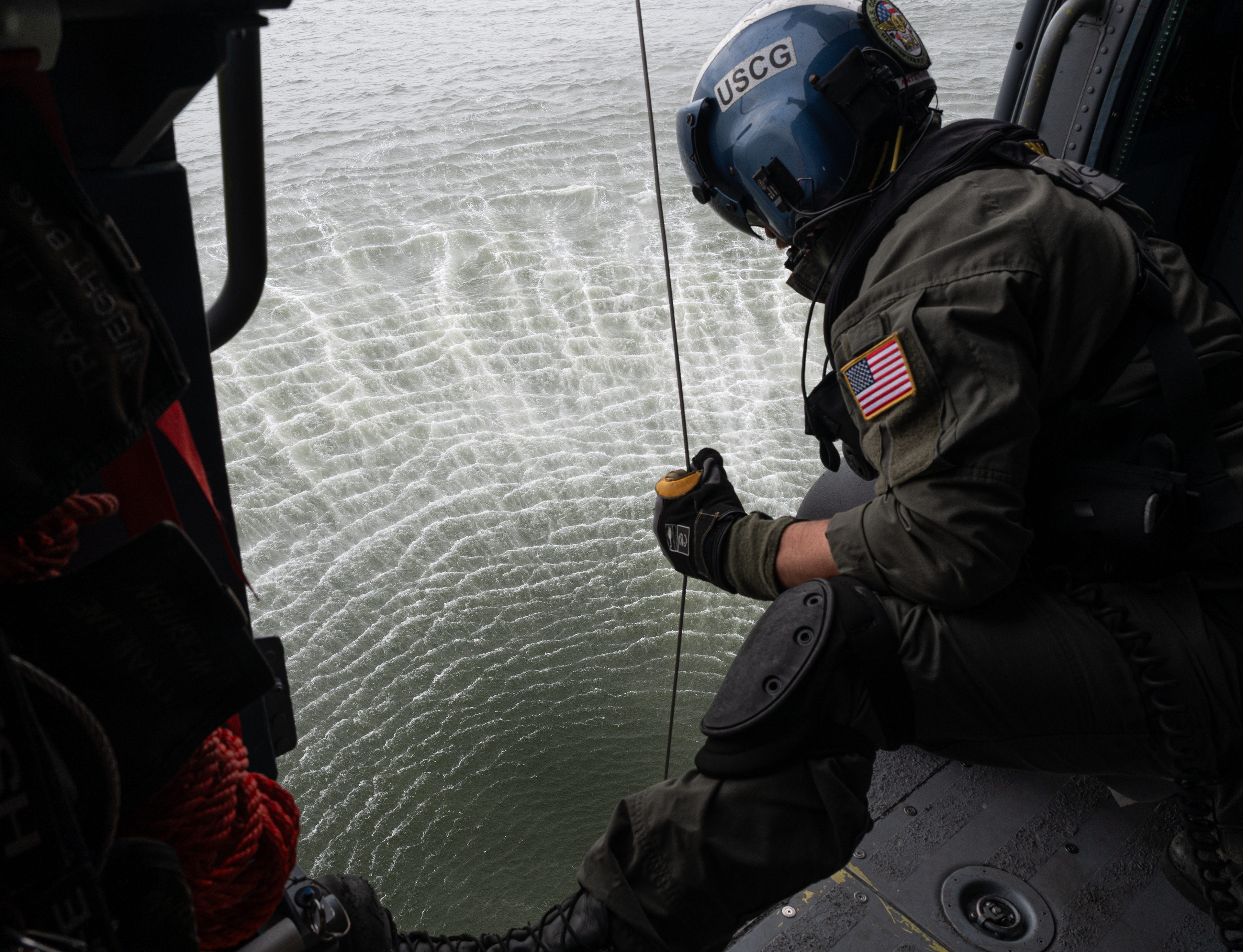 A U.S. Coast Guard aviation maintenance technician assigned to Air Station Elizabeth City, pulls the rescue back into the MH-60T Jayhawk helicopter during a search-and-rescue exercise over Cape Hatteras, North Carolina, Aug. 28, 2025. The exercise promoted mission readiness while demonstrating aircraft operations, emergency response and security procedures.