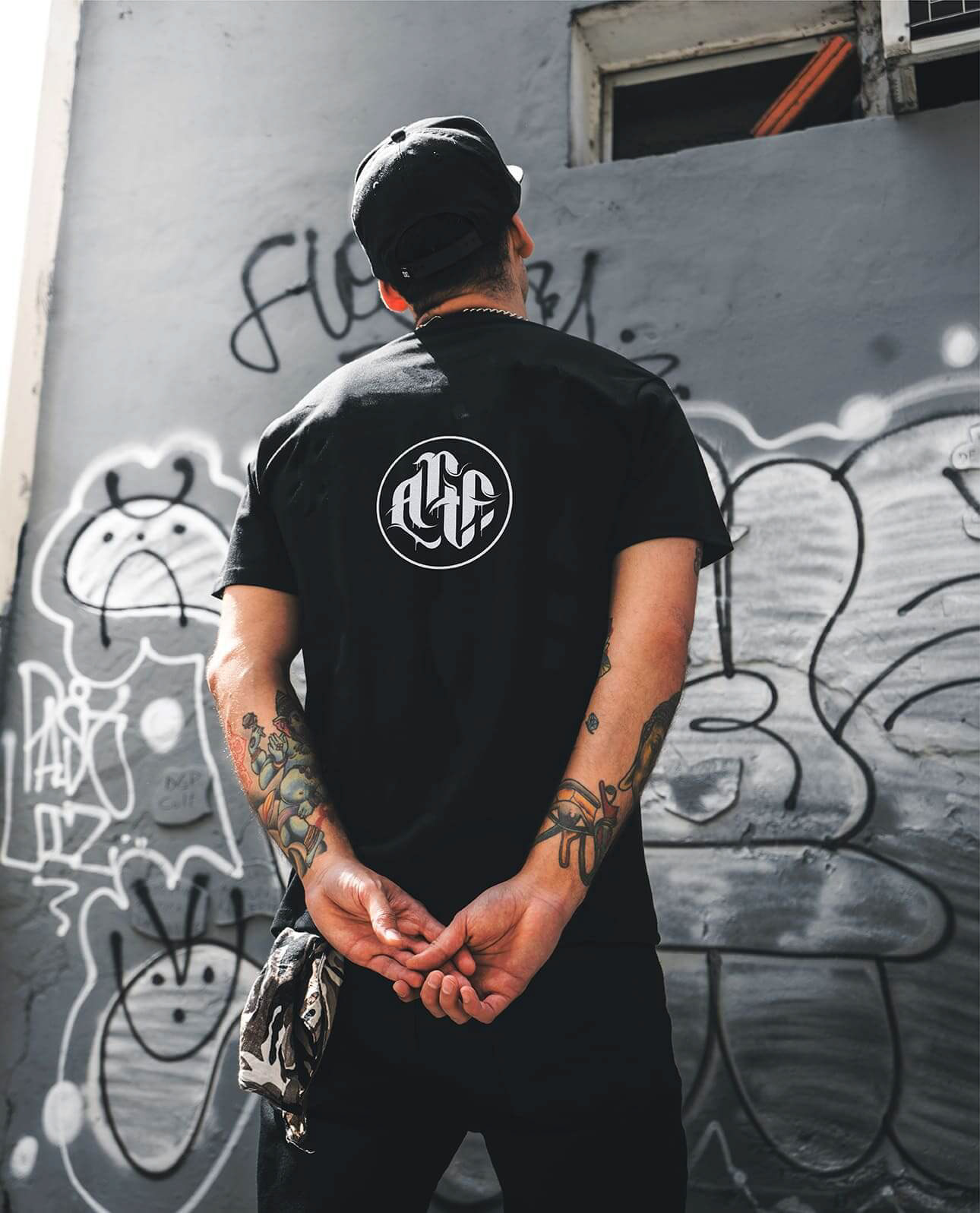 Homem jovem de costas olhando para um muro grafitado com uma camiseta preto contendo o símbolo Artficina em branco