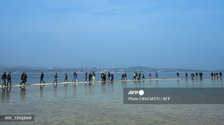 People walk towards the small island of San Biagio, off Manerba del Garda, Lake Garda, on February 21, 2023, where the water level dropped to its lowest in 30 years during the winter months. A lack of snow on the surrounding mountain peaks, the absence of rain for the past six weeks, and mild temperatures - this explosive cocktail has caused the water level of Italy's largest lake to drop to its lowest in 30 years during the winter months. (Photo by Piero CRUCIATTI / AFP)