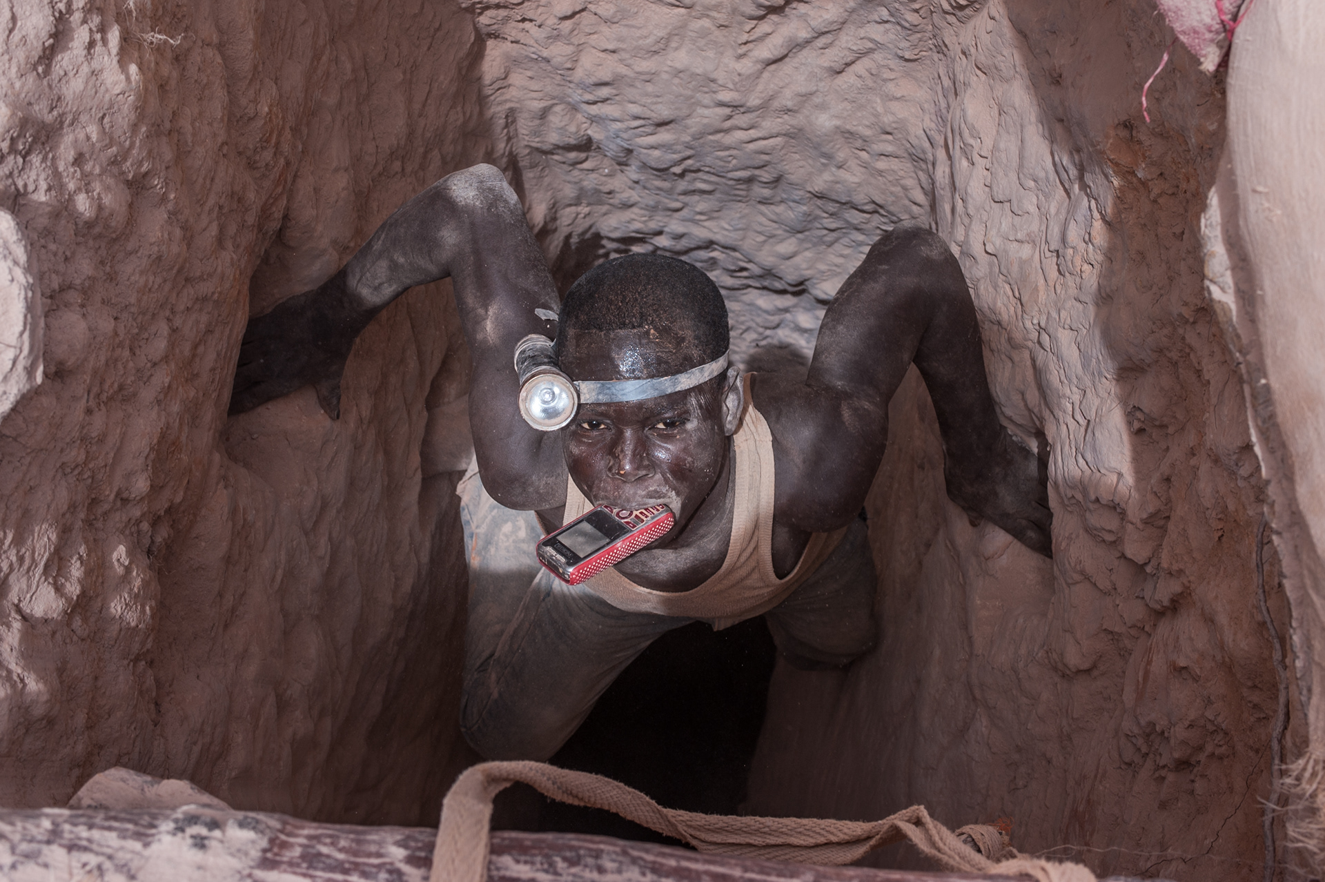 Nobsin, Burkina Faso - 13 May 2014: a young man wearing a forehead torch holds his mobile phone in his mouth as he descends in a 25 meters mine shaft. In the around 700-800 artisanal sites around the country, there’s no health and safety, no hard hats or harnesses, no protective masks, no lights and no decent ventilation system.