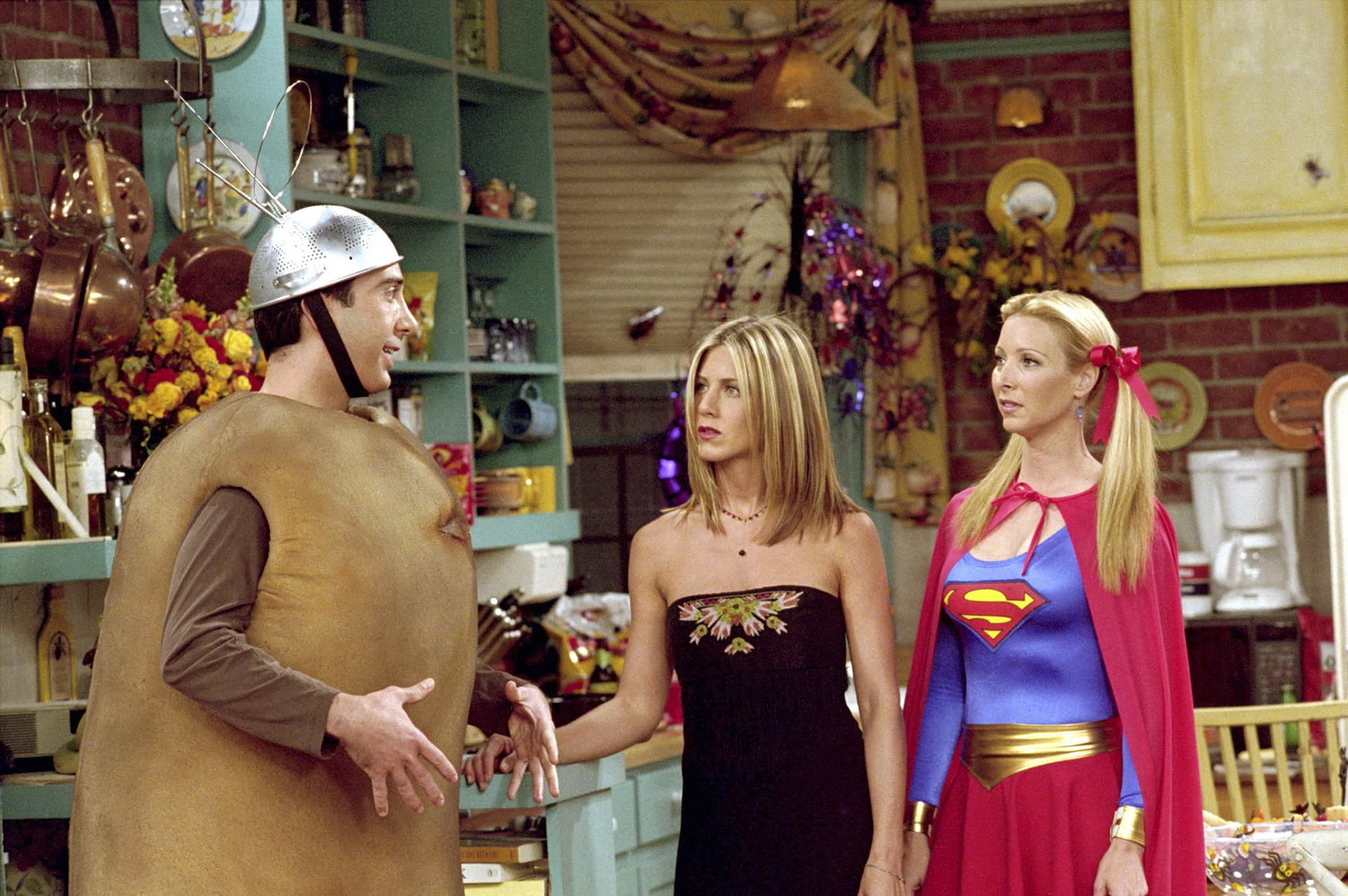 Scene from Friends. Ross is dressed as a potato with a collindar on his head, Rachel in a black dress, and Pheobe is dressed as Super Girl
