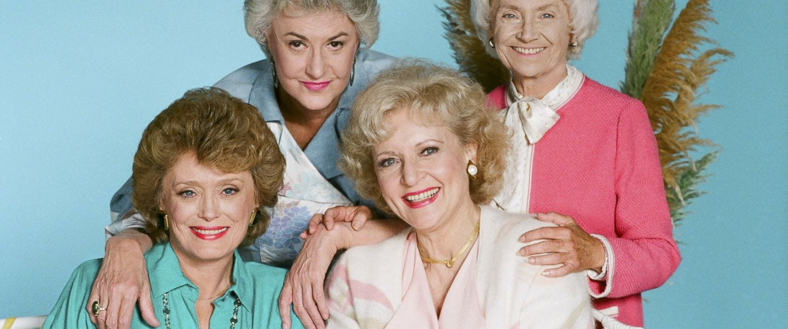 The cast of the Golden Girls - Blanche, Dorothy, Blanche and Sophia smiling at camera against blue backdrop