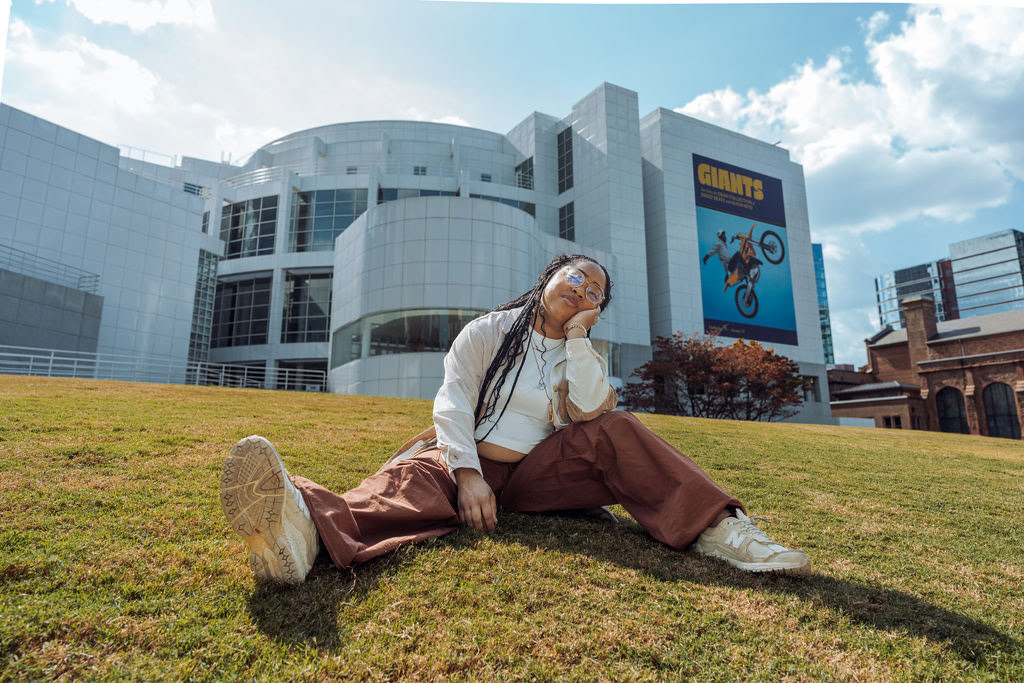 Image Description: Photograph of Sierra King, a Black Woman with braids, wearing a cream, brown and salmon colored button down shirt and rust colored cargo pants. She is sitting on the front of lawn of The High Museum of Art. She is resting her head on her hand.