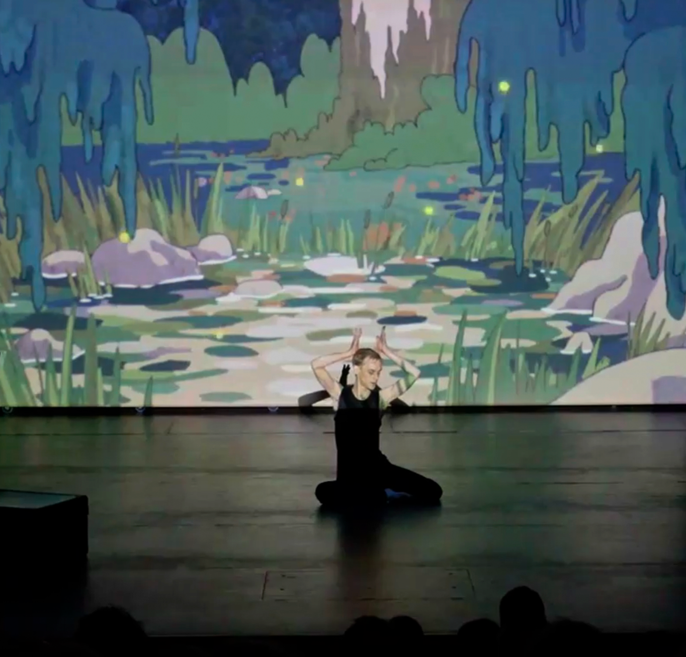 Dancer sitting on stage in front of an illustration of a pond projected behind him