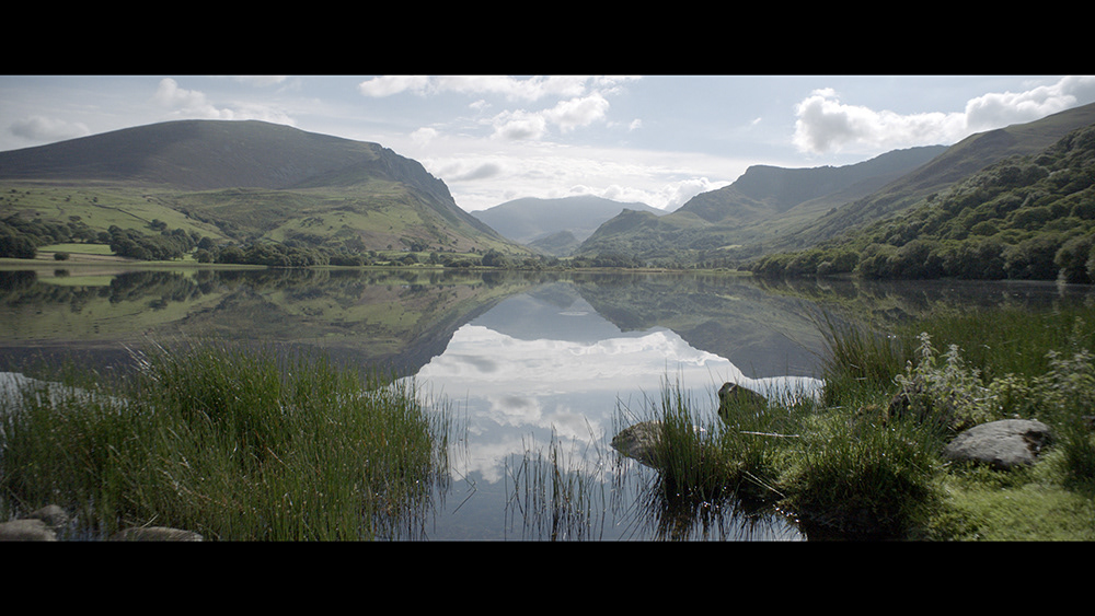 Beautiful picture of a landscape with a light blue sky, clouds and mountains reflecting in the water of a lake and green vegetation in the foreground