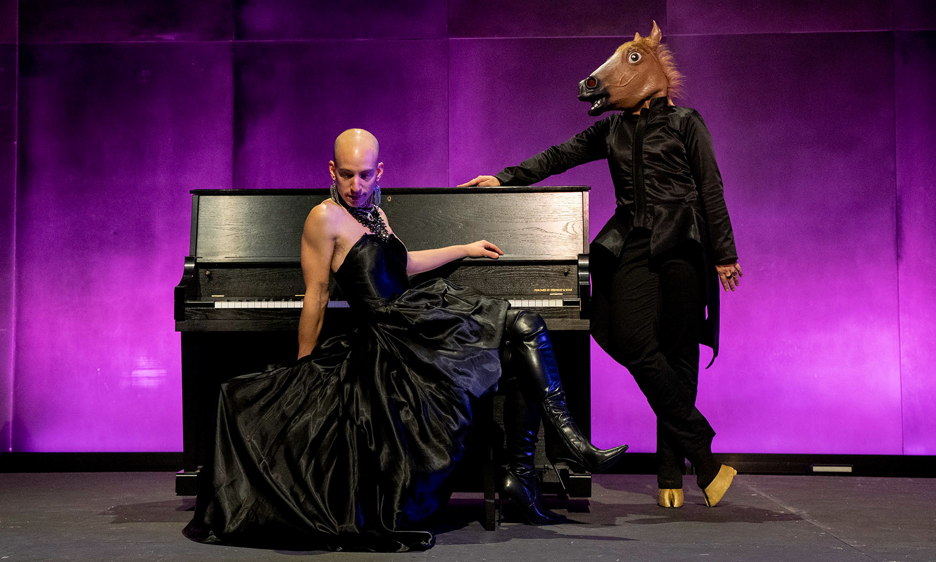 A person sits at a piano in a formal black dress and looks down beside another person in a formal pant suit and tails wearing a horse mask