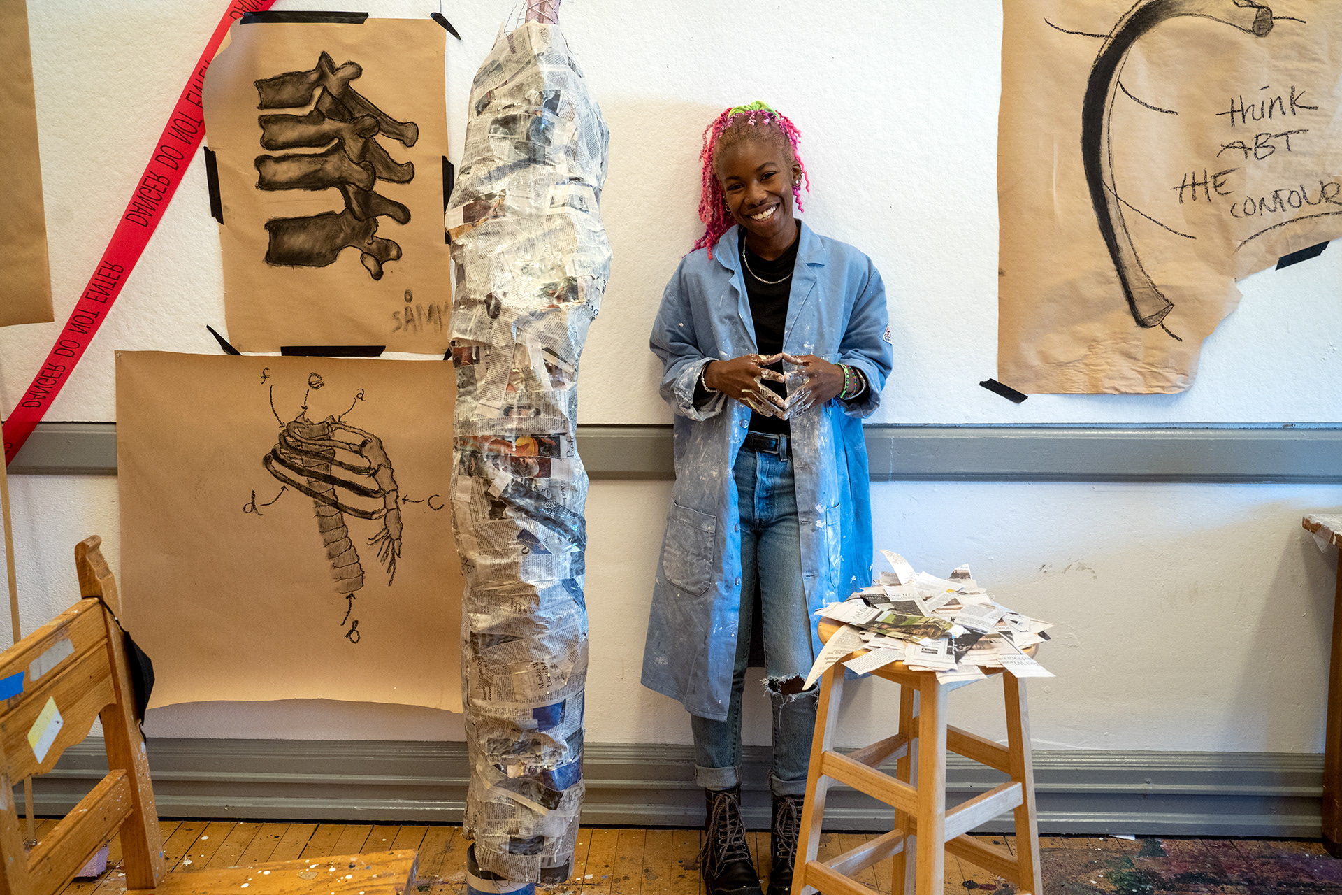 An artist stands against a wall beside a large object covered in newspaper and surrounded by sketches.