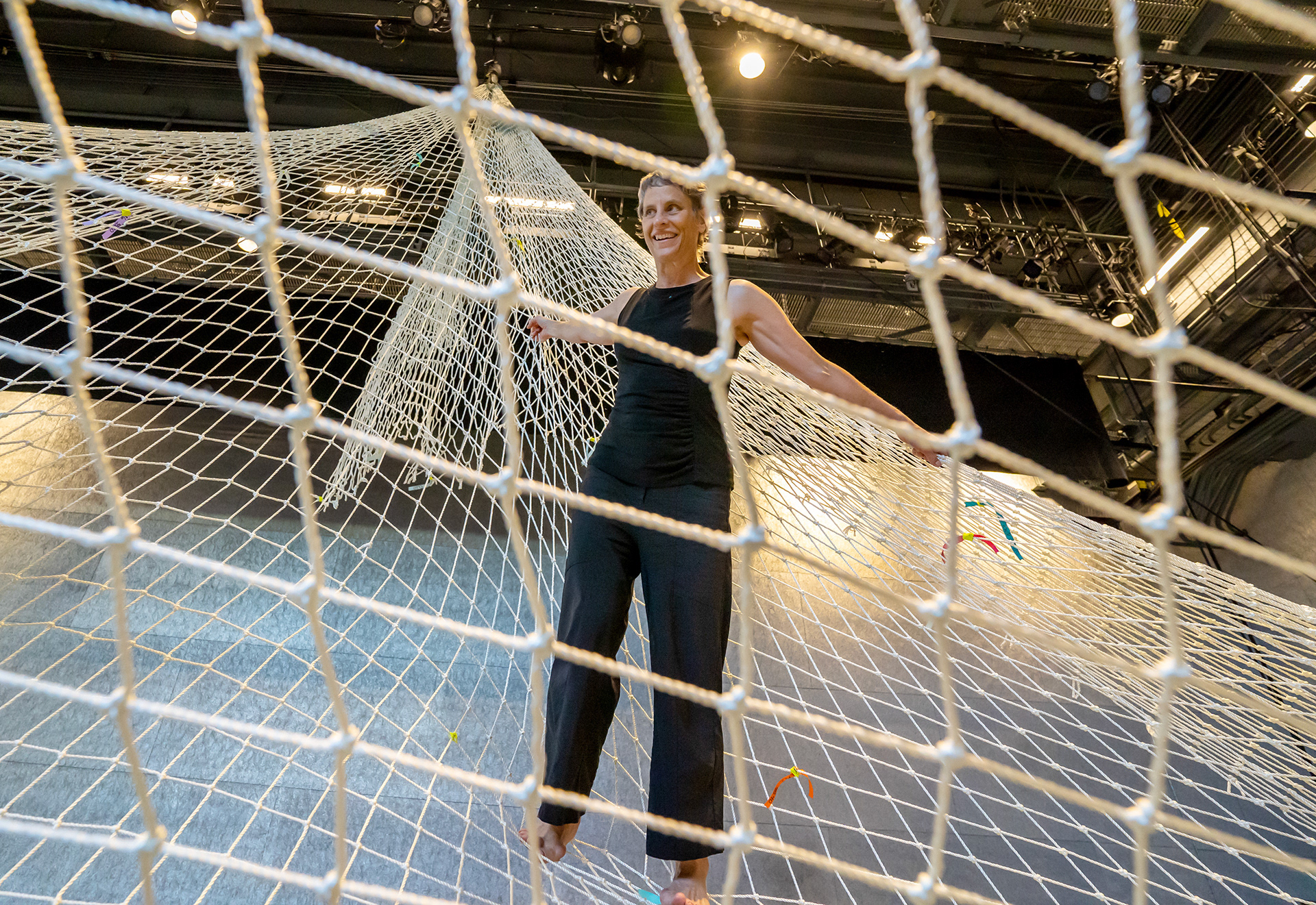 A person stands in a large net hanging from a high ceiling in a theatrical space