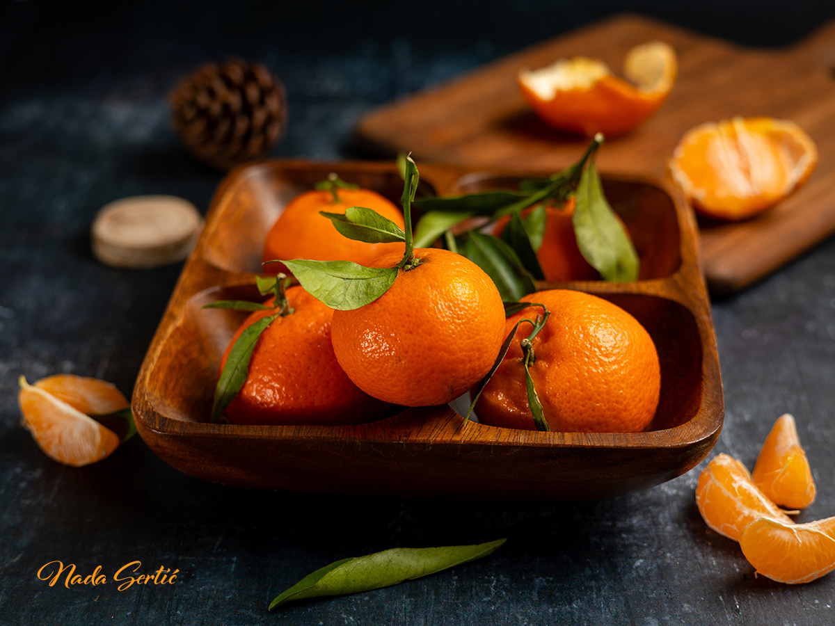 Clementines in wooden bowl on dark background with pieces scattered around.