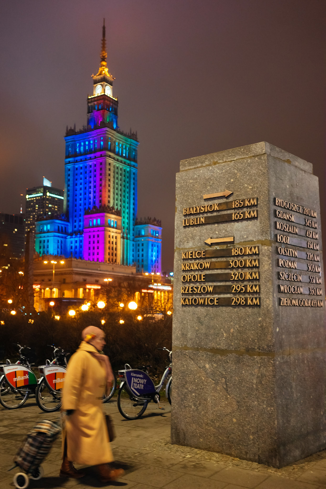Varşova şehir merkezinden bir gece manzarası. Arka planda her akşam belirli bir süre rengarenk aydınlatılan Kültür ve Bilim Sarayı, önde ise Polonya'nın çeşitli şehirlerine olan mesafelerin belirtildiği anıt görülüyor.