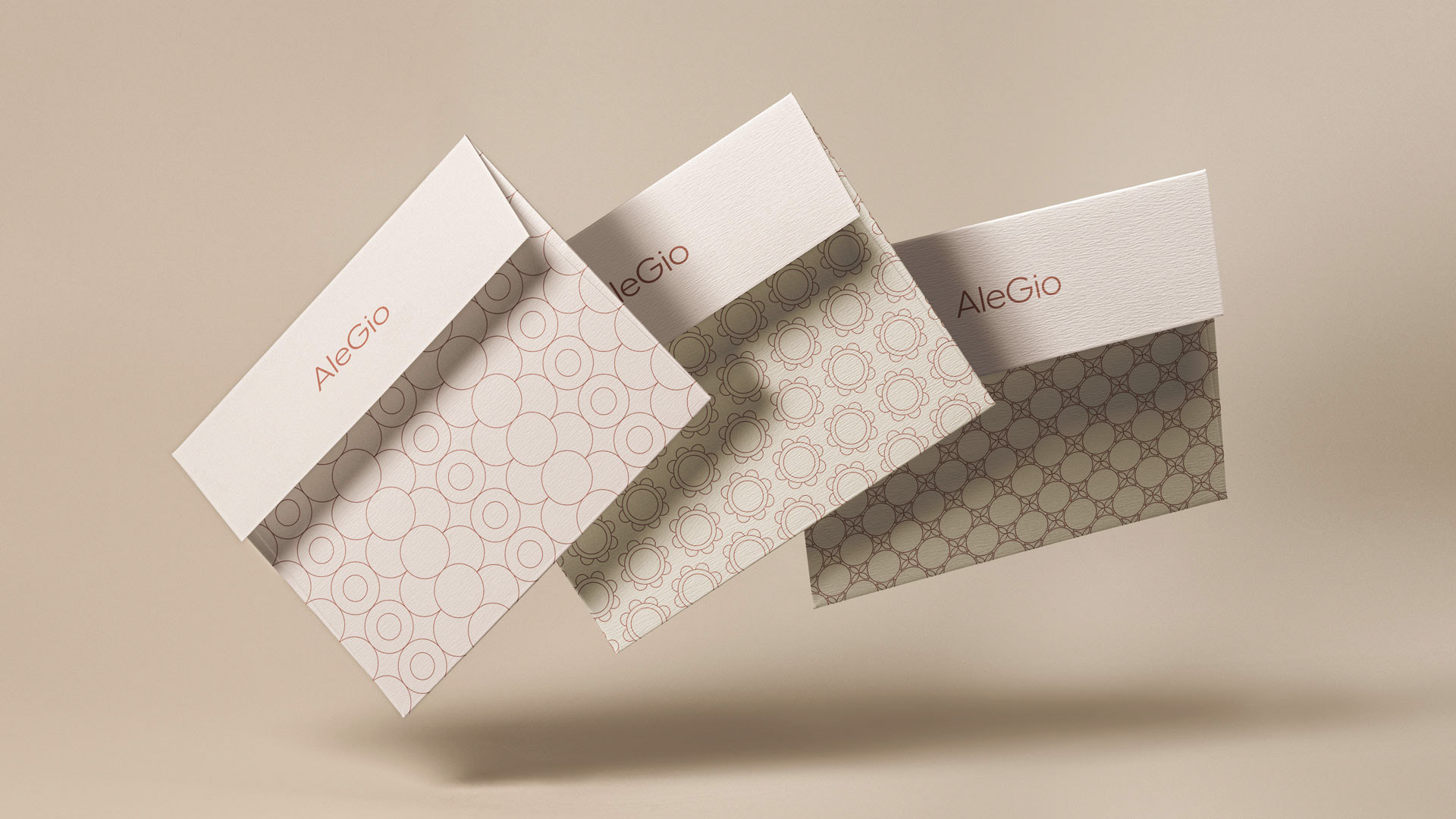 Three AleGio envelopes in the air, made of beige paper. The envelopes are adorned with the logotype and each has a unique pattern, all in bronze foil.
