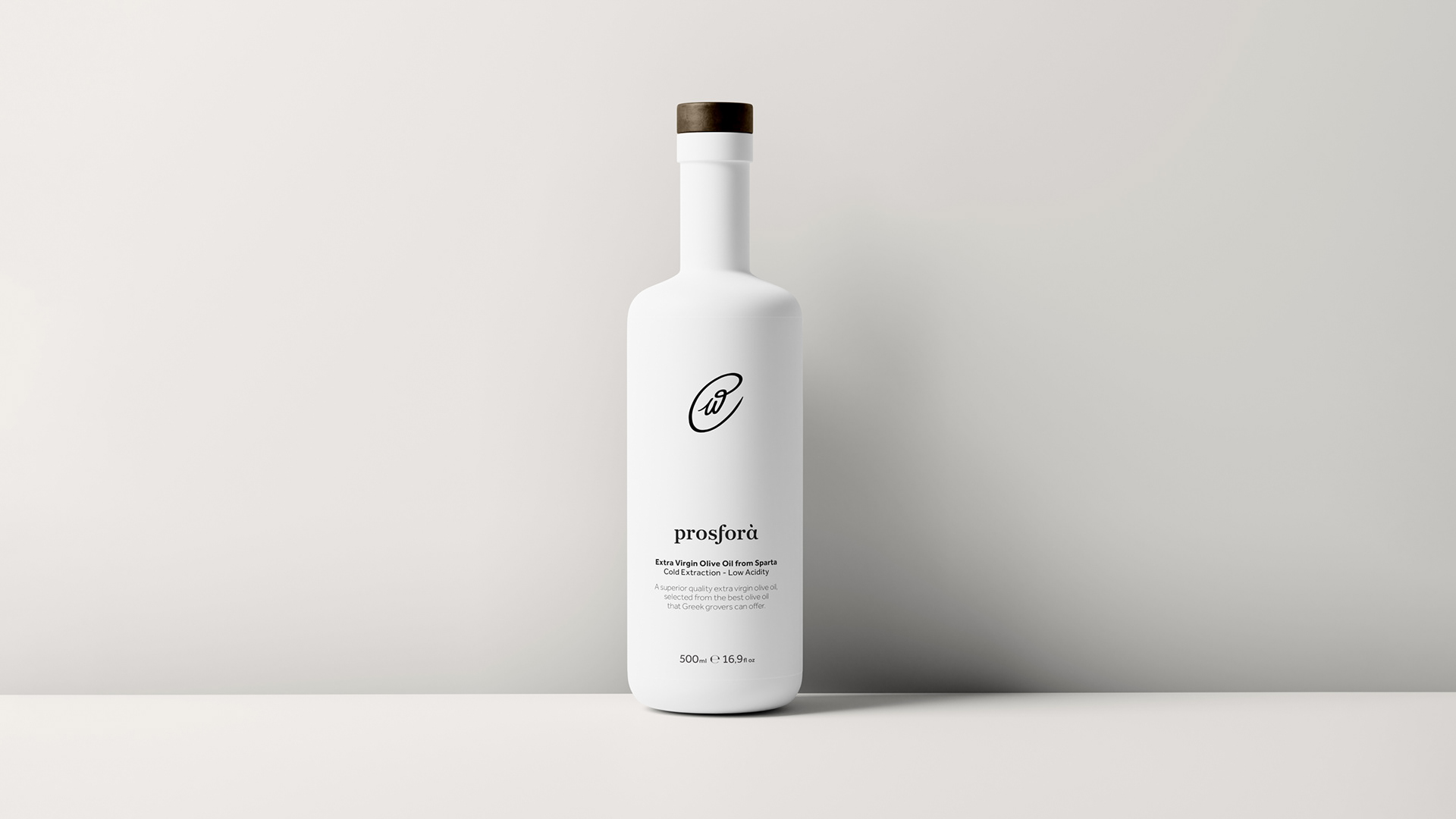 Prosfora olive oil white bottle standing against a wall. The bottle features the prosfora logotype that resembles an olive at its center, while typography is in the bottom part of the bottle.