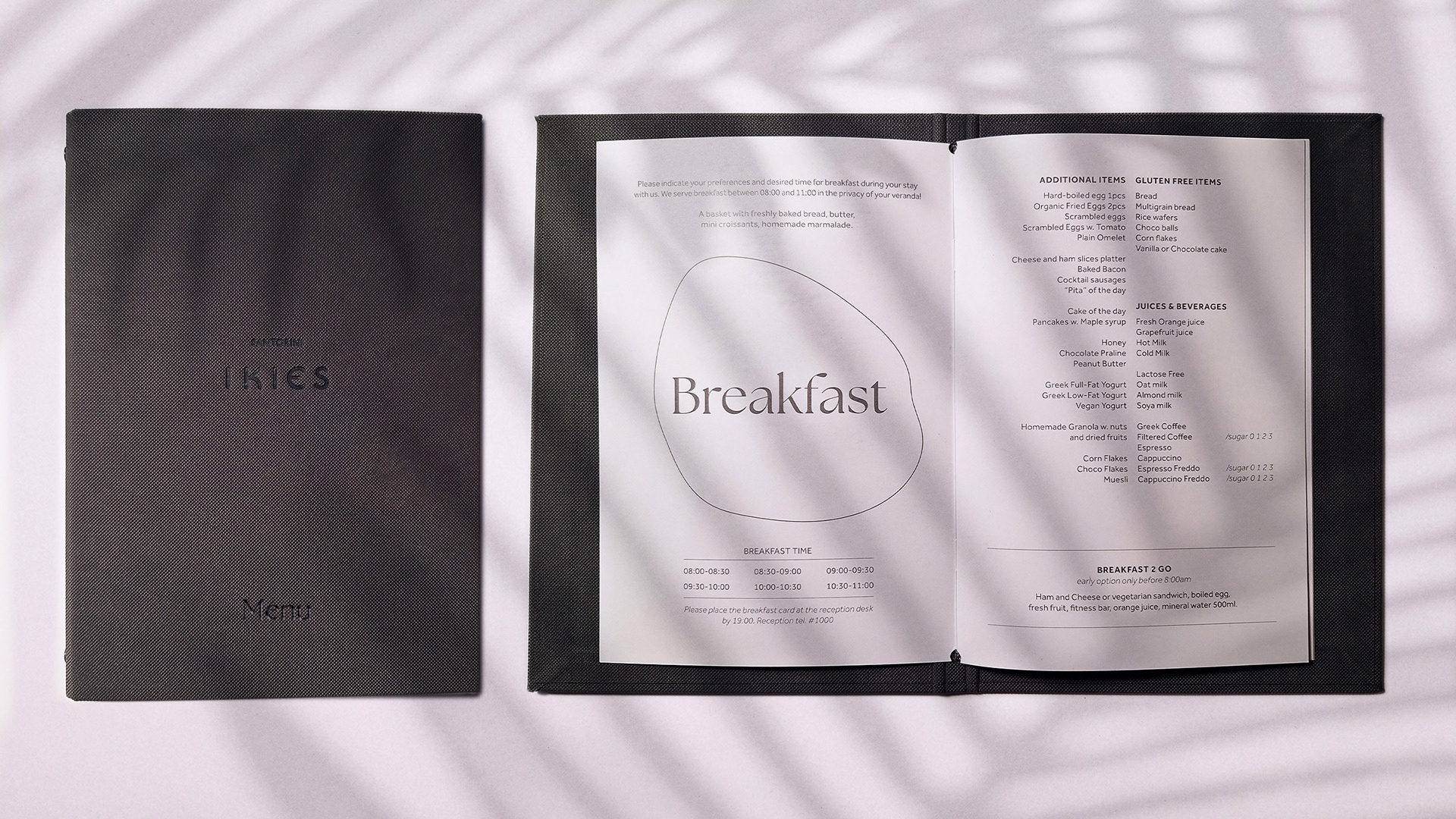 IKIES Santorini boutique hotel menu design showing the PU leather cover with embossed logo and title and a second menu is opened at the breakfast section showcasing the layout. The image is taken in the shadow of a plant.