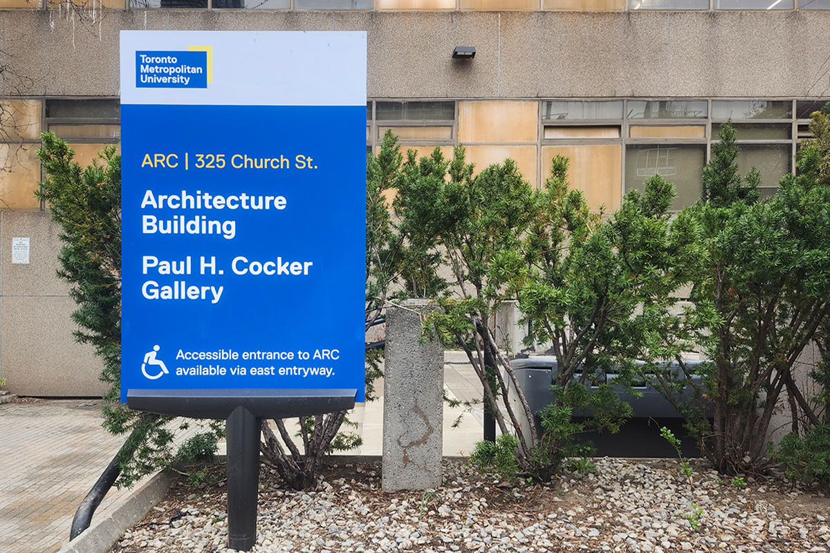 Photograph of an exterior building ID which includes additional information to direct people to an alternate accessible entrance. 