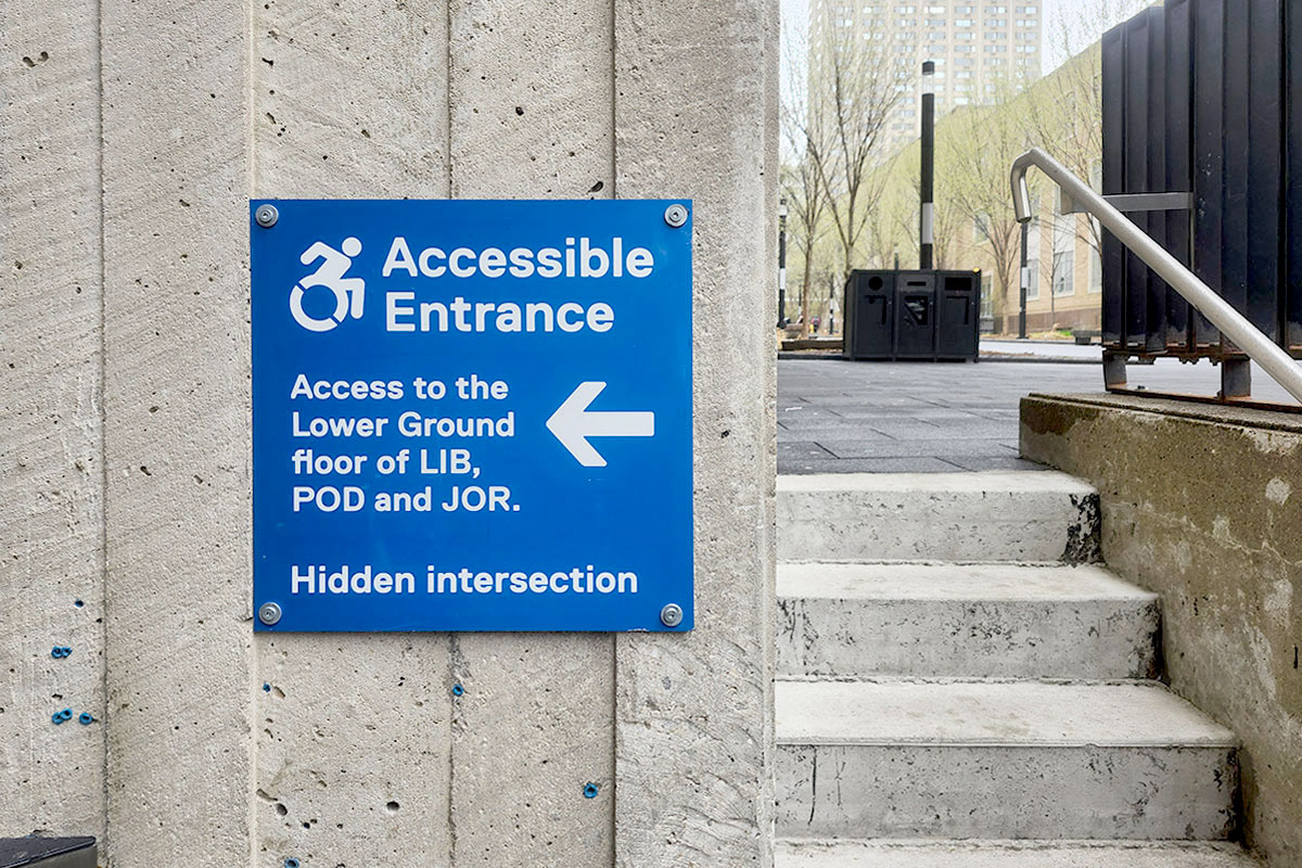 Photograph of an exterior sign that helps direct users to a hidden accessible entrance.