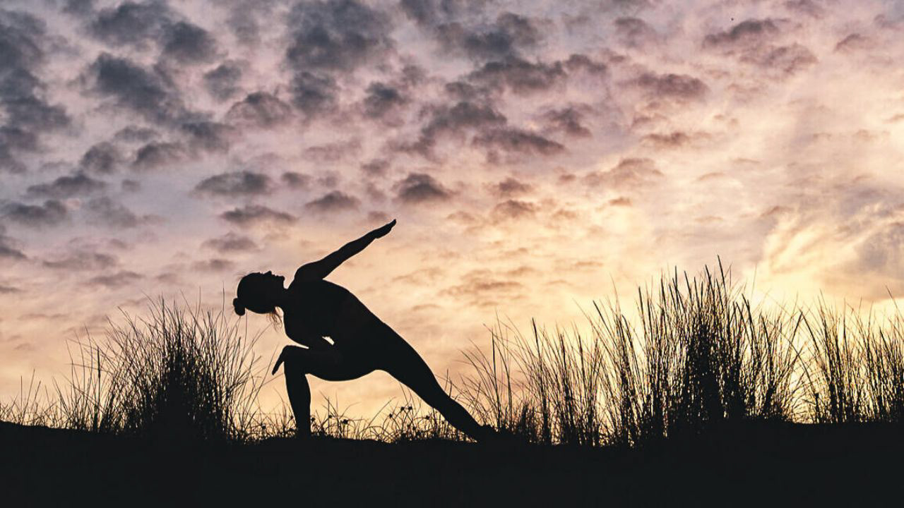 A silhouetted person practices a yoga pose on a a grassy dune at sunset, showcasing a peaceful, outdoor wellness session in nature
