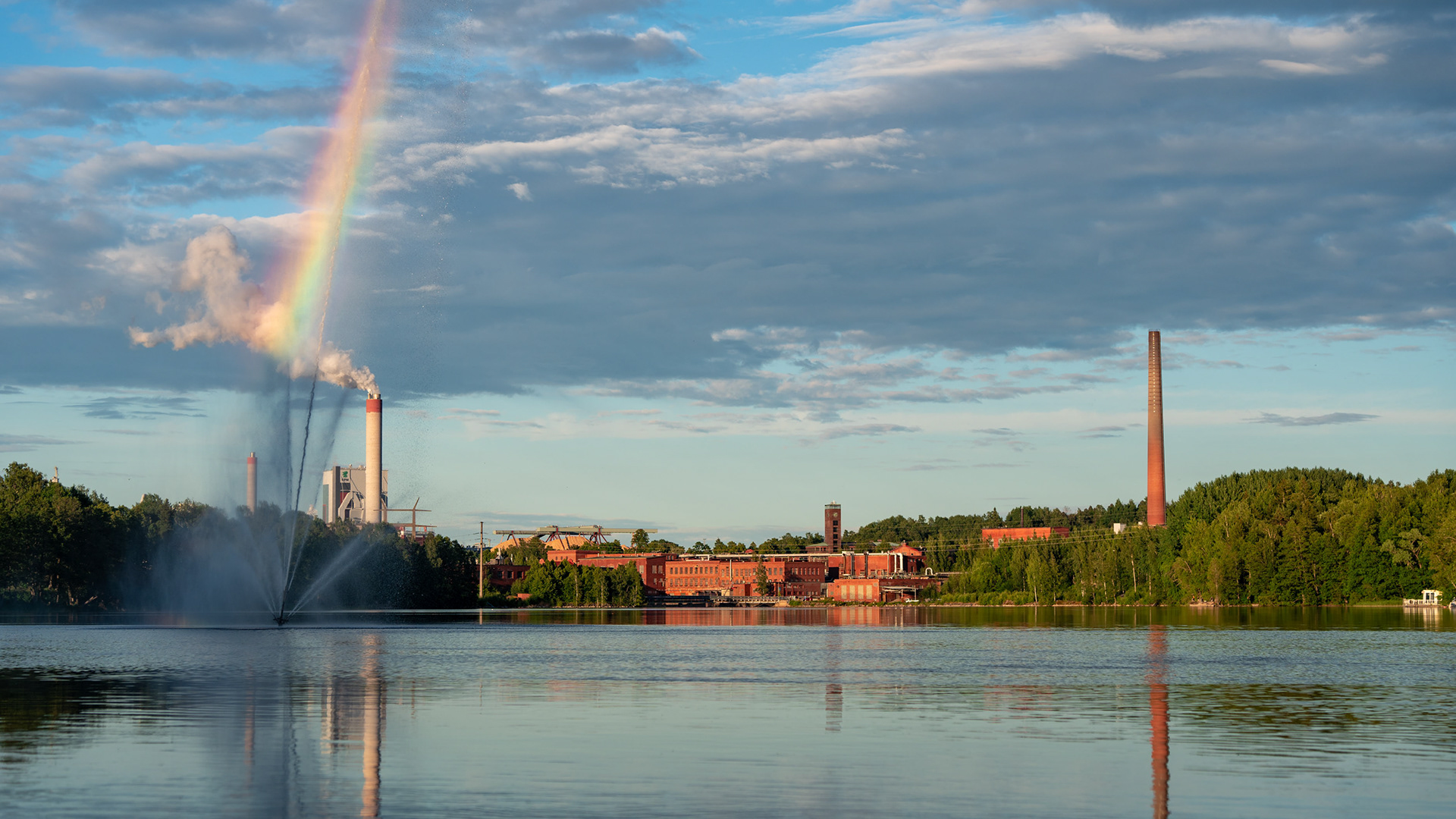 Näkymä Kuusan sillan kohdalta joelle Kymintehtaalle päin. Etualalla rantapuiston suihkulähde. Taustalla Kymin vanha tehdasalue sekä nykyinen UPM Kymin paperitehdas. (6.2024)
