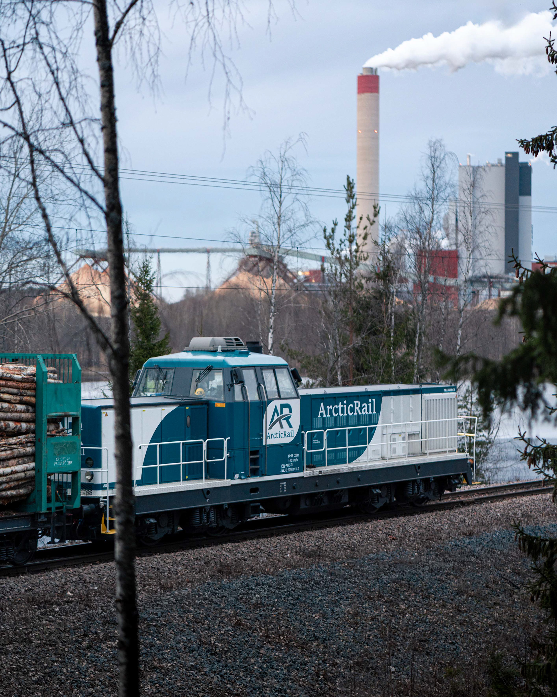 ArcticRailin Dr16 -dieselveturin vetämä tukkijuna matkalla Kouvolan lajittelusta UPM Kymin tehtaalle Kuusankoskelle. Kuvassa juna saapumassa Rapakosken sillalle ja taustalla Kymijoki sekä UPM Kymi.  (21.3.2026)