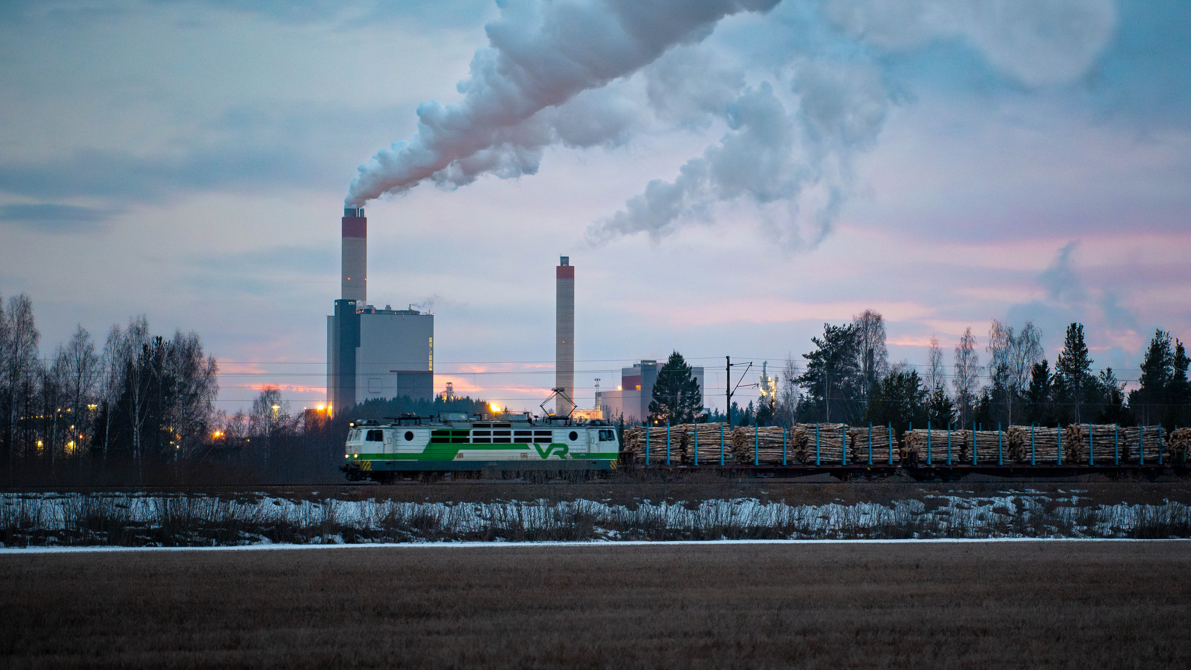 Sr1-sähköveturin vetämä täysi tukkijuna matkalla Pieksämäeltä Kouvolan lajitteluun. Taustalla UPM-Kymin paperi- ja sellutehdas. (20.3.2025)