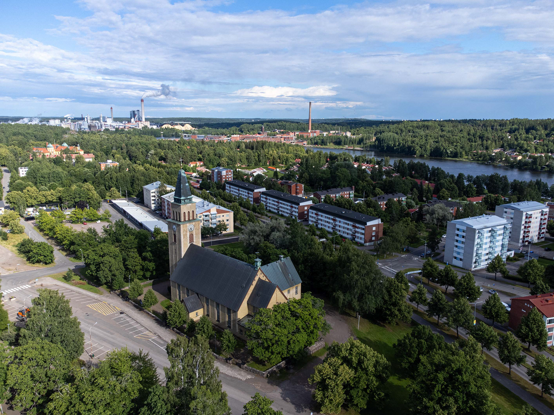 Kuusankosken kirkko ja kirkonpuistoa. Taustalla Kymin vanha tehdasalue ja nykyinen UPM Kymi (7.2022)